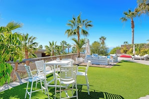 Une terrasse extérieure avec des meubles blancs, des palmiers et un ciel d’un bleu éclatant.