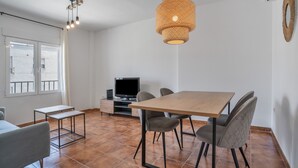 Un salón moderno con una mesa de comedor de madera, sillas grises, un televisor de pantalla plana sobre un mueble bajo, una mesita auxiliar y un amplio ventanal con cortinas.