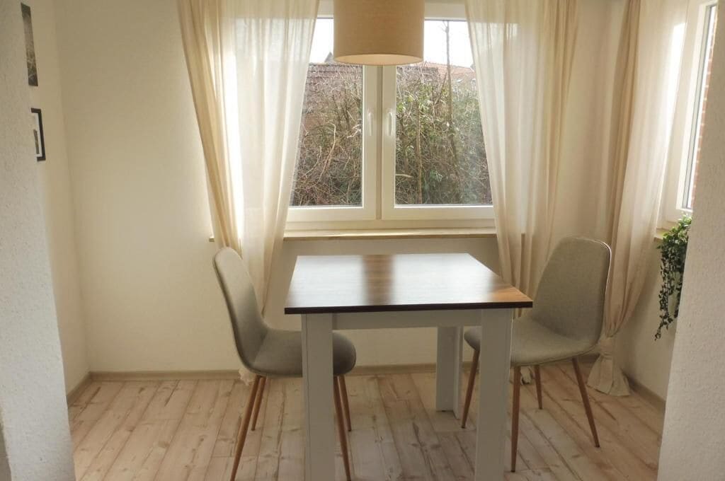 A room with a table and two chairs, a window with curtains, and a plant.