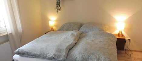 A bed with a white mattress and two pillows, two bedside lamps, a small plant on a shelf, and a framed picture on the wall.