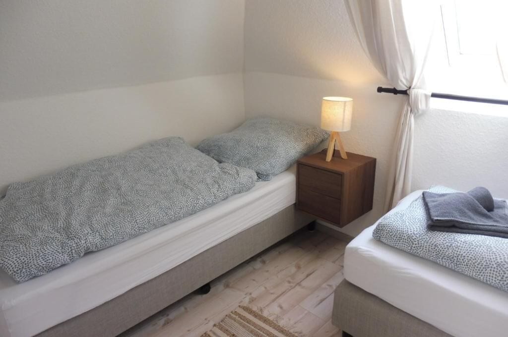 Une petite chambre avec un lit simple, une table de chevet en bois, une lampe et une fenêtre avec des rideaux.