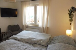 Habitación de hotel con una cama, una silla, un televisor montado en la pared, una ventana con cortinas y una lámpara al lado.