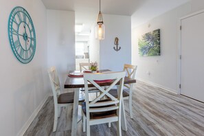A dining area with a wooden table, white chairs, a large clock, and a hanging light fixture. - CNB10- West Beach Jewel! Amazing View! Enjoy The Beach & Lagoon! (Gulf Shores)