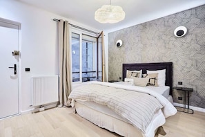 A bedroom with a large bed, a radiator, a window with curtains, a wall with a textured pattern, and a hanging light fixture. - Superb Studio in the Center of Paris/Opera (Paris)