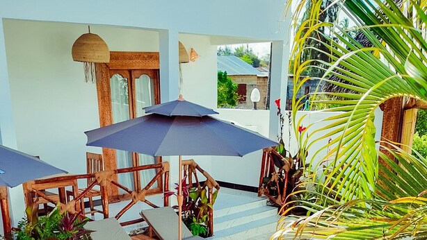 Zona de piscina con sillones de jardín y sombrillas, rodeada de plantas y un pequeño edificio.