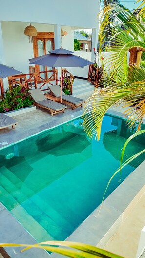 A pool area with lounge chairs and umbrellas, surrounded by plants and a small building.