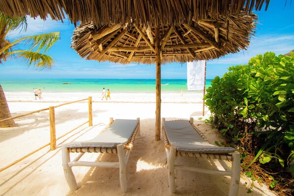 A beach with two lounge chairs under a thatched roof, a sandy path, and a clear view of the ocean.