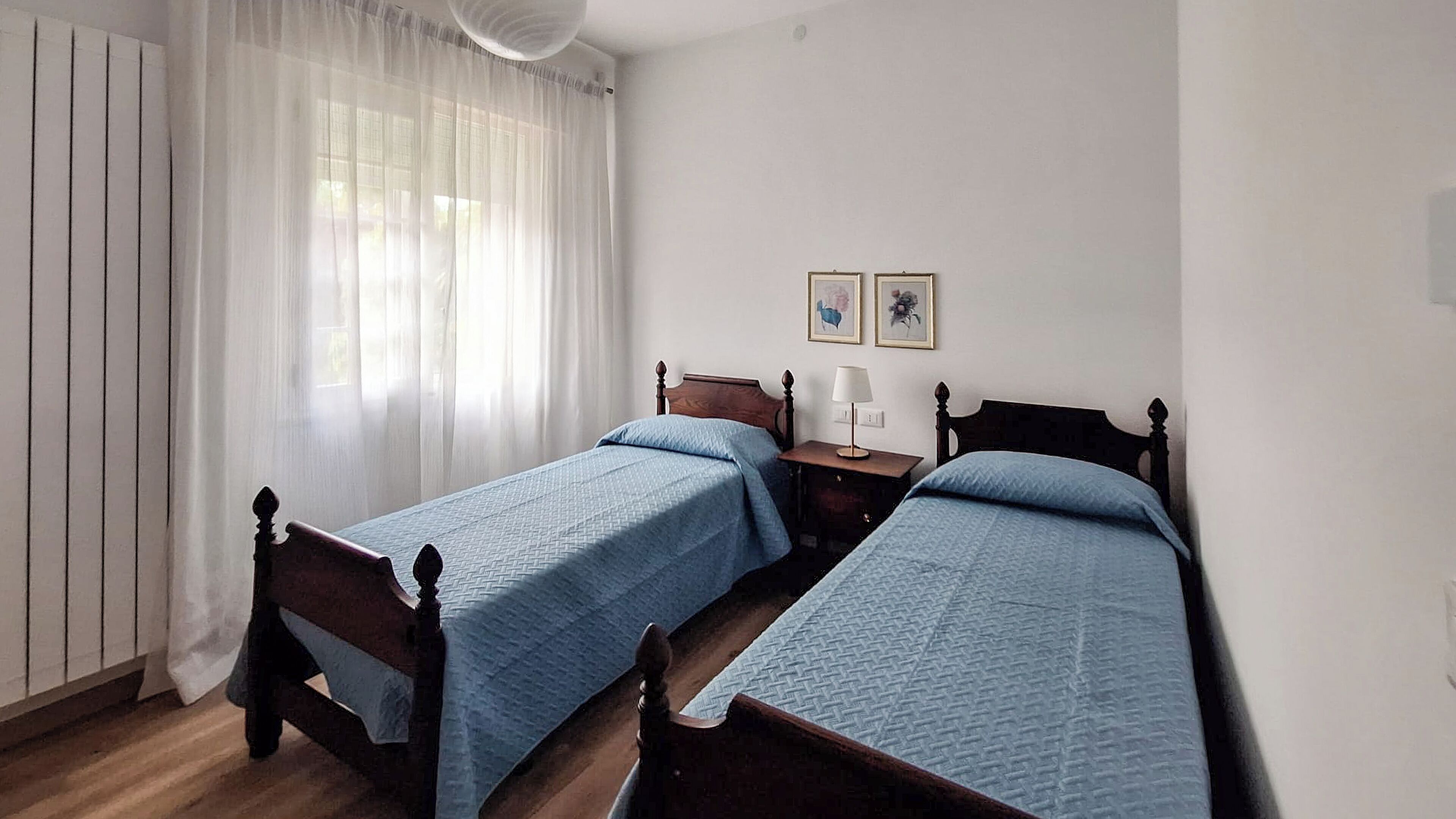 Une chambre à coucher avec deux lits en bois, une table de chevet avec une lampe, une fenêtre avec des rideaux légers et deux tableaux encadrés accrochés au mur.
