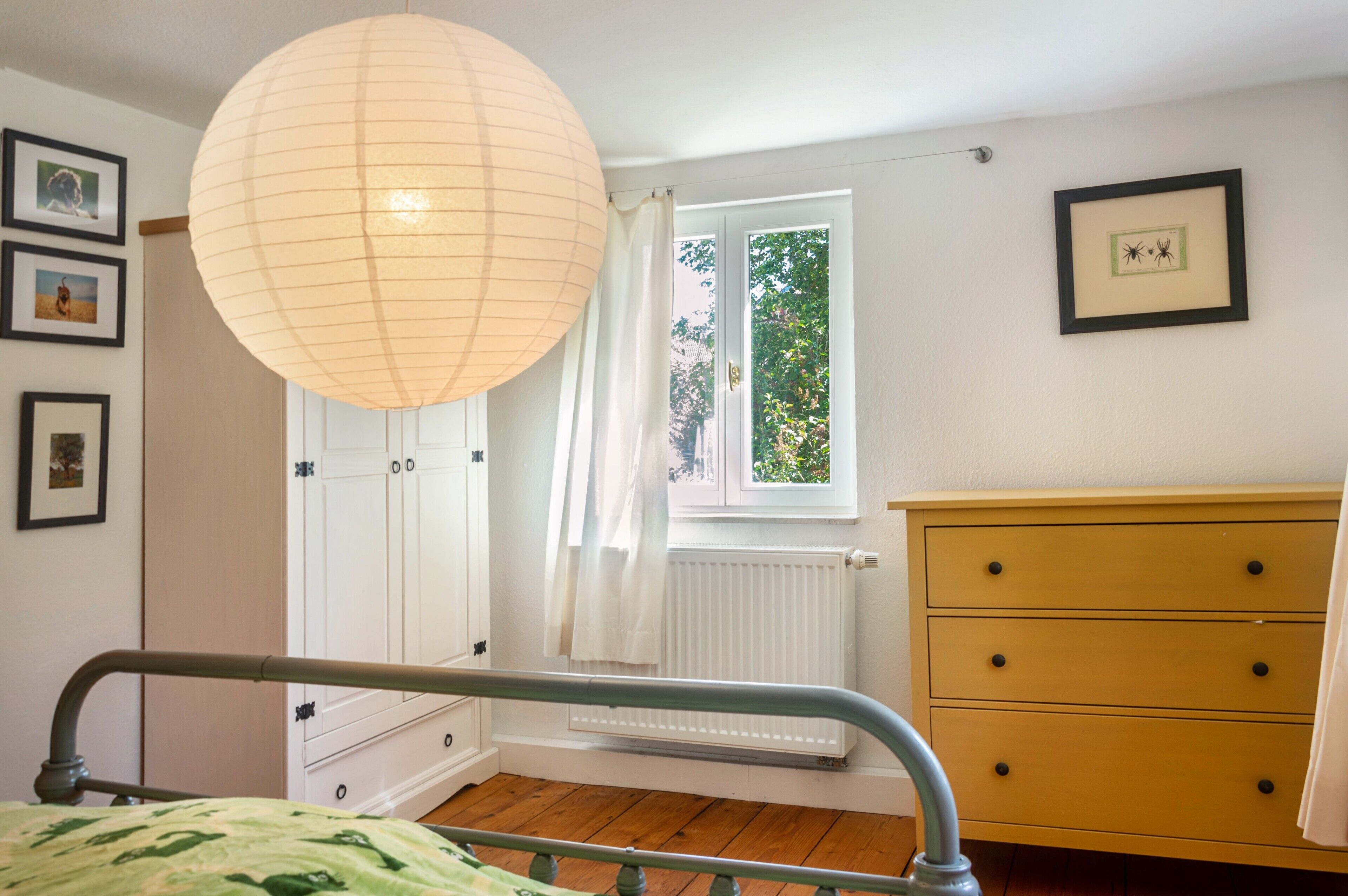 A bedroom with a bed, a dresser, a radiator, a window with curtains, and framed pictures on the wall.