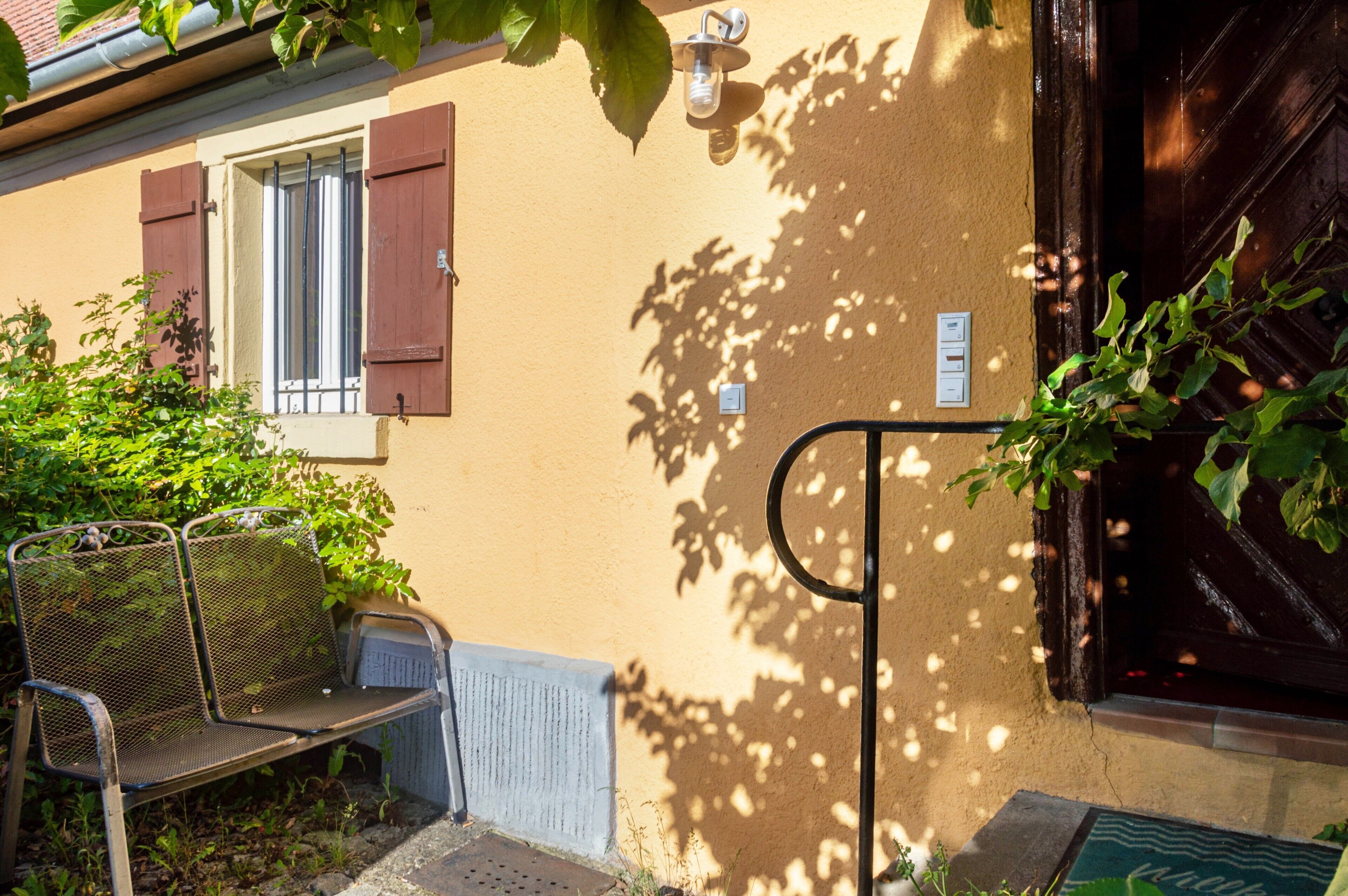 Un banc devant un bâtiment doté d’une porte et d’une fenêtre, d’un interrupteur mural et de l’ombre d’un arbre.
