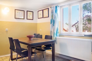 Un coin repas avec une table et des chaises, des tableaux encadrés au mur, une fenêtre avec des rideaux et un radiateur.