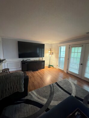 Ein Wohnzimmer mit Flachbildfernseher, einem dunkelgrauen Sofa, einem gemusterten Teppich und einer Stehlampe.