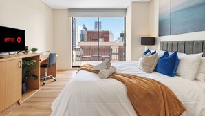 Una habitación de hotel moderna con una cama grande, un escritorio con una computadora, un televisor mostrando el logotipo de Netflix y un balcón con vistas a la ciudad.
