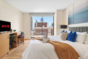 Ein modernes Hotelzimmer mit einem großen Bett, einem Schreibtisch mit Computer, einem Fernseher mit Netflix-Logo und einem Balkon mit Blick auf die Stadt.