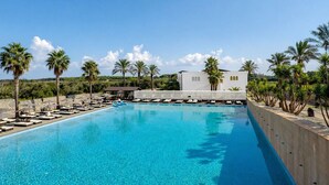 Una piscina azul cristalina con sillones de descanso y palmeras, rodeada por un edificio blanco con ventanas arqueadas.
