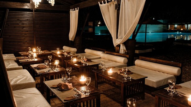 Un espacio exterior para comer con mesas de madera, asientos blancos acolchados y un ambiente iluminado por velas.