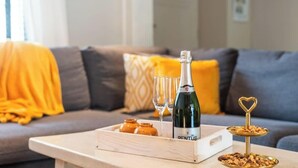 Una botella de vino, copas y una bandeja con aperitivos sobre una mesa de madera en una sala de estar con sofá y cojines.