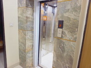 An elevator with a marble wall, a digital display showing the number 2, and a sign with emergency instructions. - Hotel Shree Krishna Inn (Ujjain)