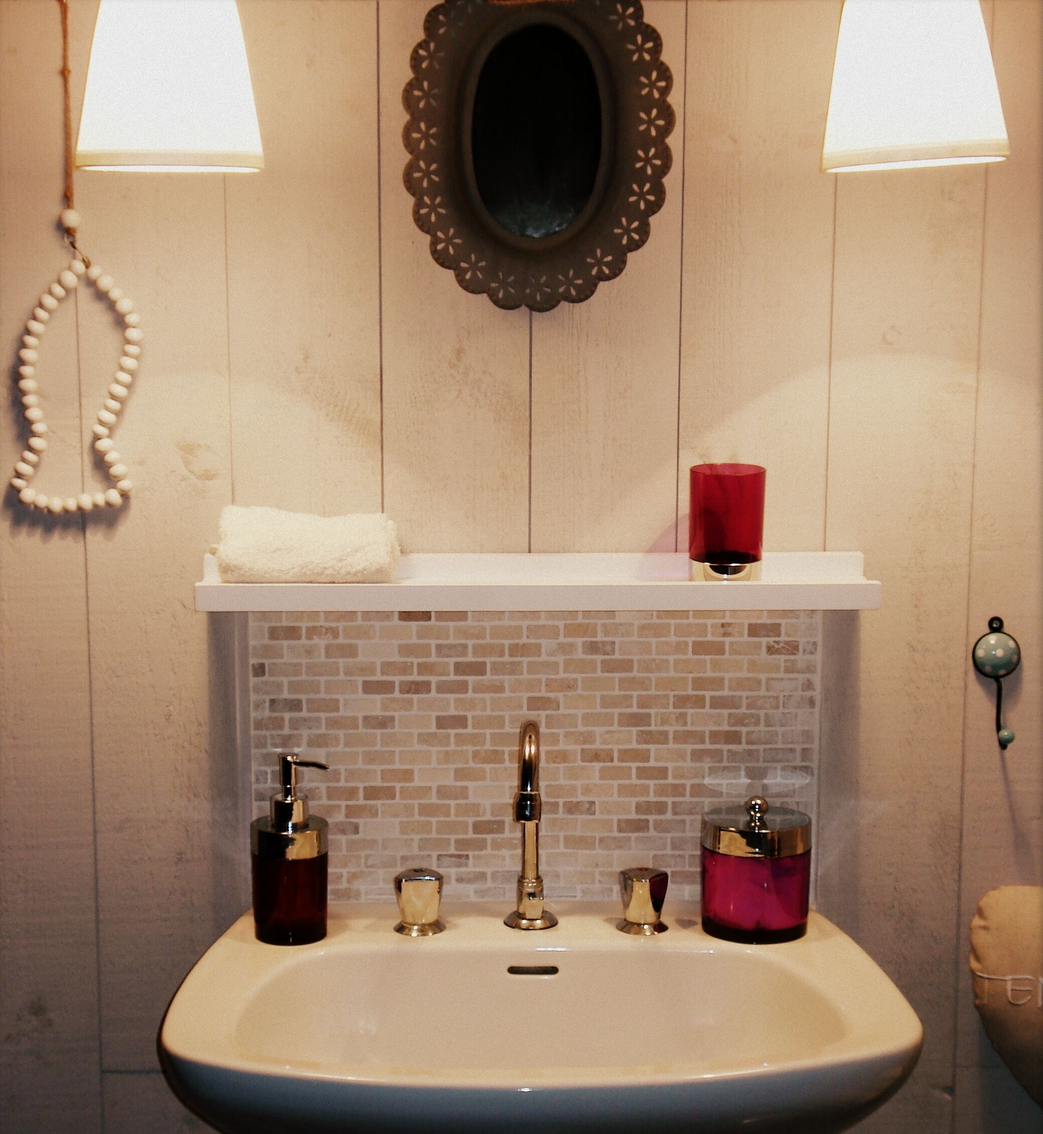 Un baño con un lavabo blanco, dos luces empotradas en la pared, un espejo y una repisa con una toalla y una vela roja.