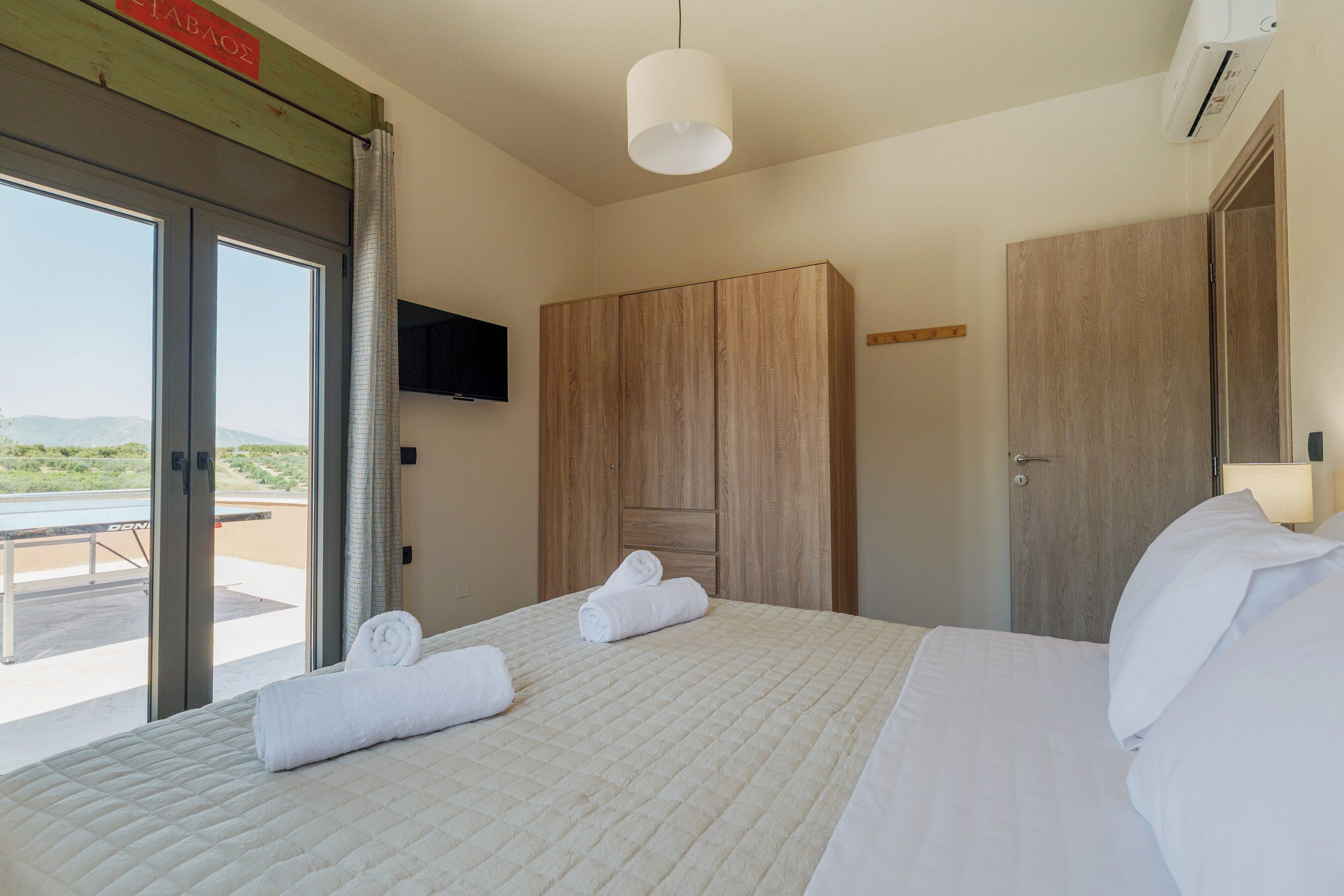 Una habitación de hotel con cama, un armario de madera, televisor montado en la pared y una puerta corrediza de vidrio que da a un espacio exterior.