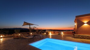 Una piscina con sillas de descanso y una mesa, rodeada por un muro de piedra y vistas al atardecer.