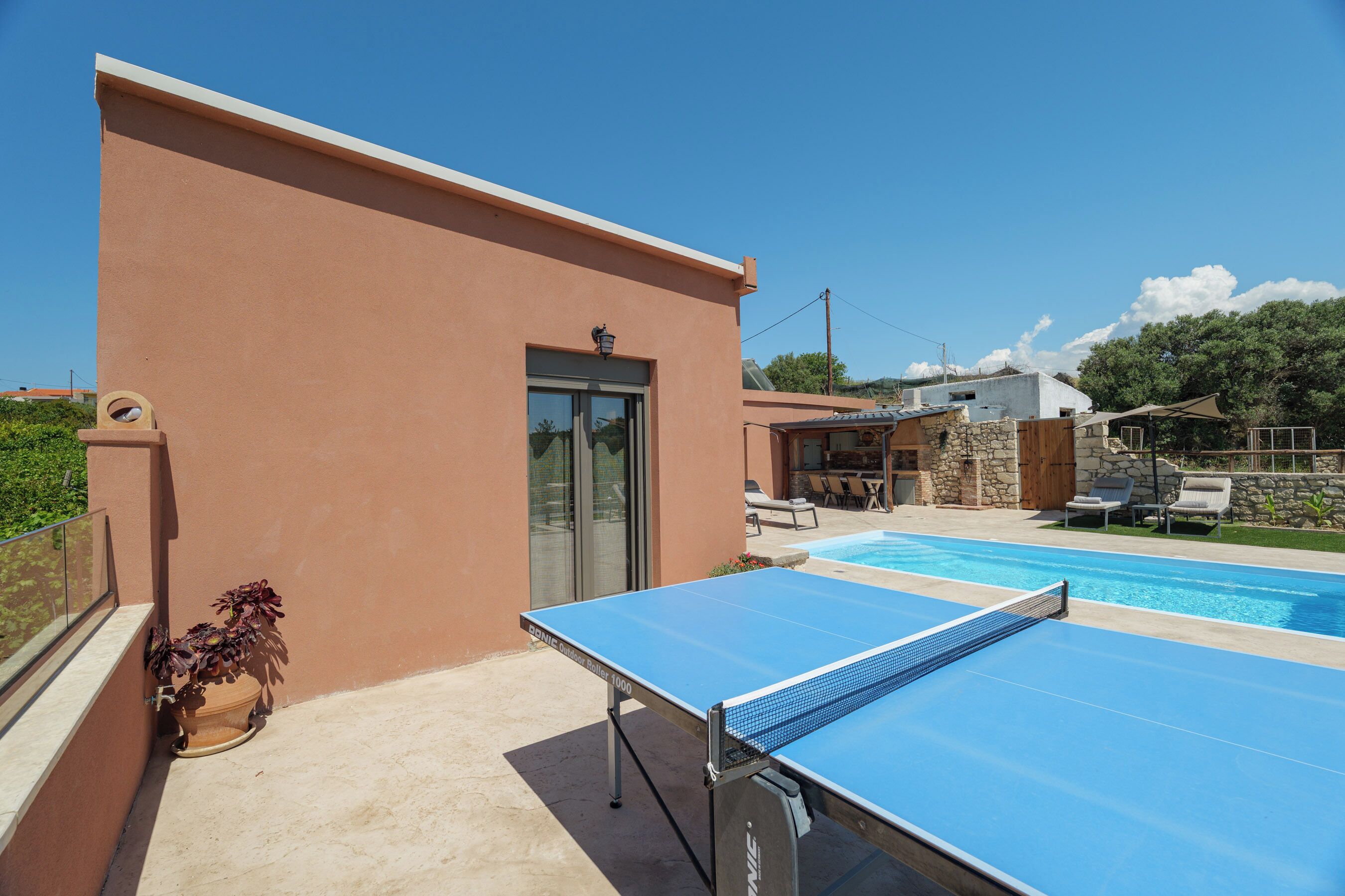 Mesa de bilhar numa área externa ensolarada, com piscina, um edifício de pedra e uma porta.