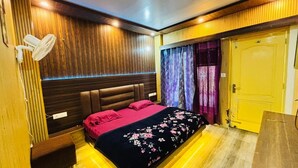 A bedroom with a bed, a bedside table, a wall-mounted fan, a door, and a curtain. - Shubh Hotel (Mussoorie)