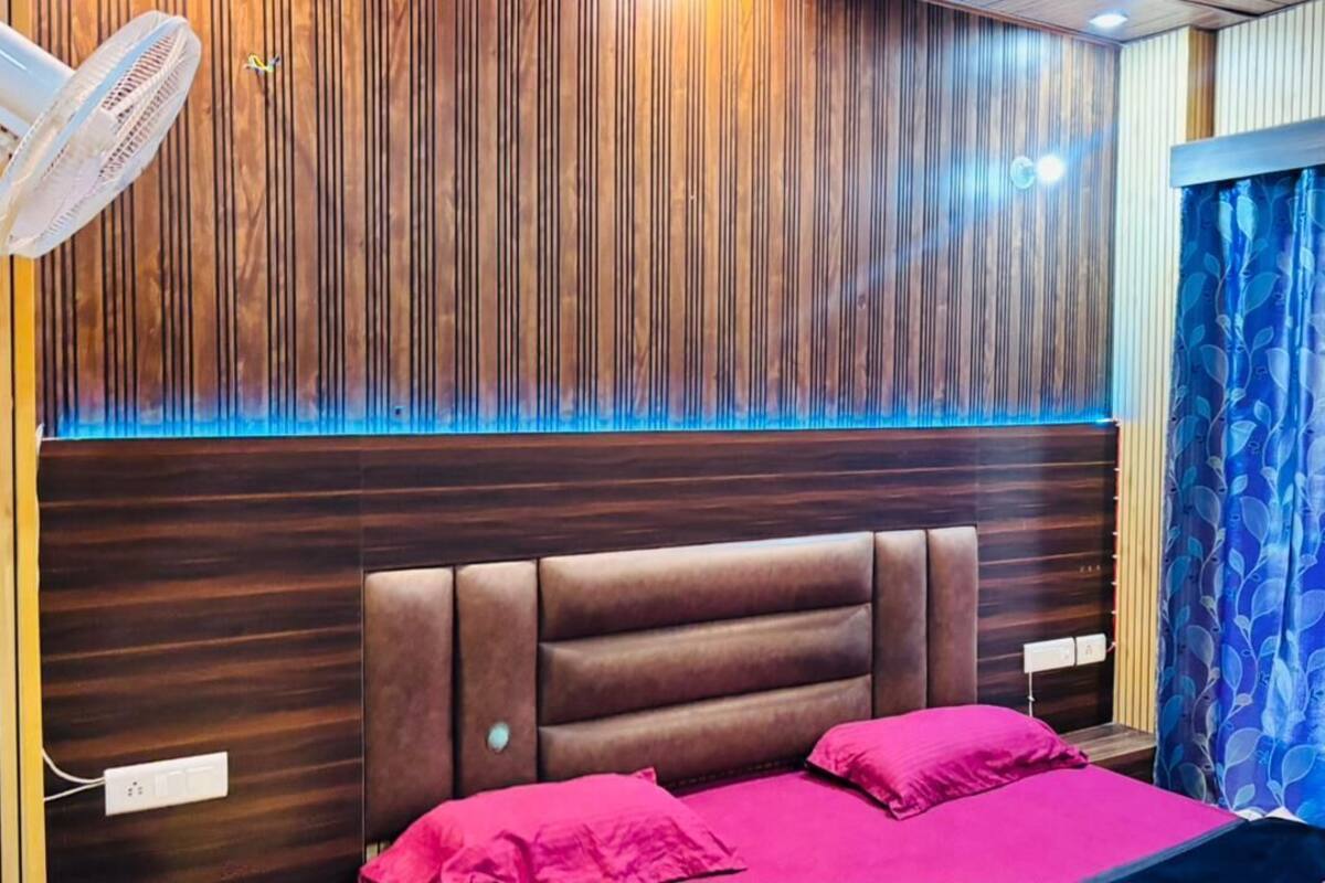 A room with a wooden panel wall, a headboard, a bed with pink bedding, and a fan.