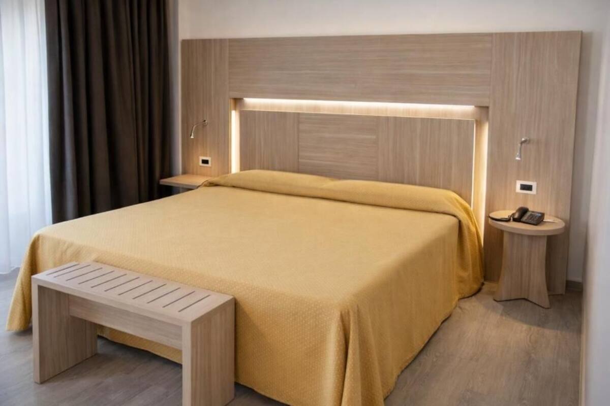 A modern hotel room with a large bed, bedside tables, a bench, and a headboard with integrated lighting.
