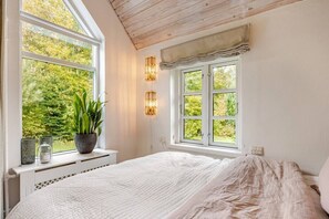 A bedroom with a large window, a bed, a potted plant, and wall-mounted lighting.