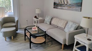 A modern living room with a sofa, armchair, glass coffee table, and a painting of sailboats.