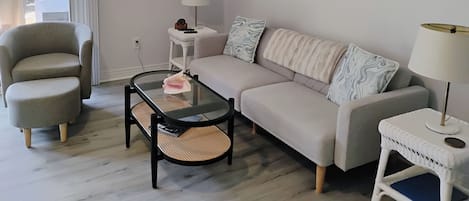 Een moderne woonkamer met een bank, fauteuil, glazen salontafel en een schilderij van zeilboten.