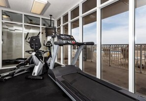 Ein Fitnessstudio mit Laufband, Crosstrainer und Ergometer, großen Fenstern und einem Balkon mit Sitzgelegenheiten.