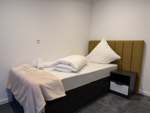 A neatly made bed with white bedding, a brown headboard, and a small bedside table with a drawer.