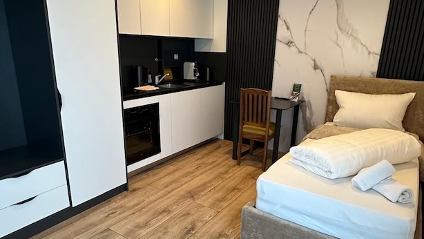 Quarto de hotel compacto com cama, pequena escrivaninha, cadeira, cozinha compacta e guarda-roupa.
