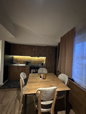 A dining area with a wooden table and chairs, a kitchen in the background, and a large window with curtains.