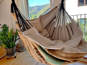 A hammock chair with a beige cushion, a potted plant, and a view of a forested hillside through a window.