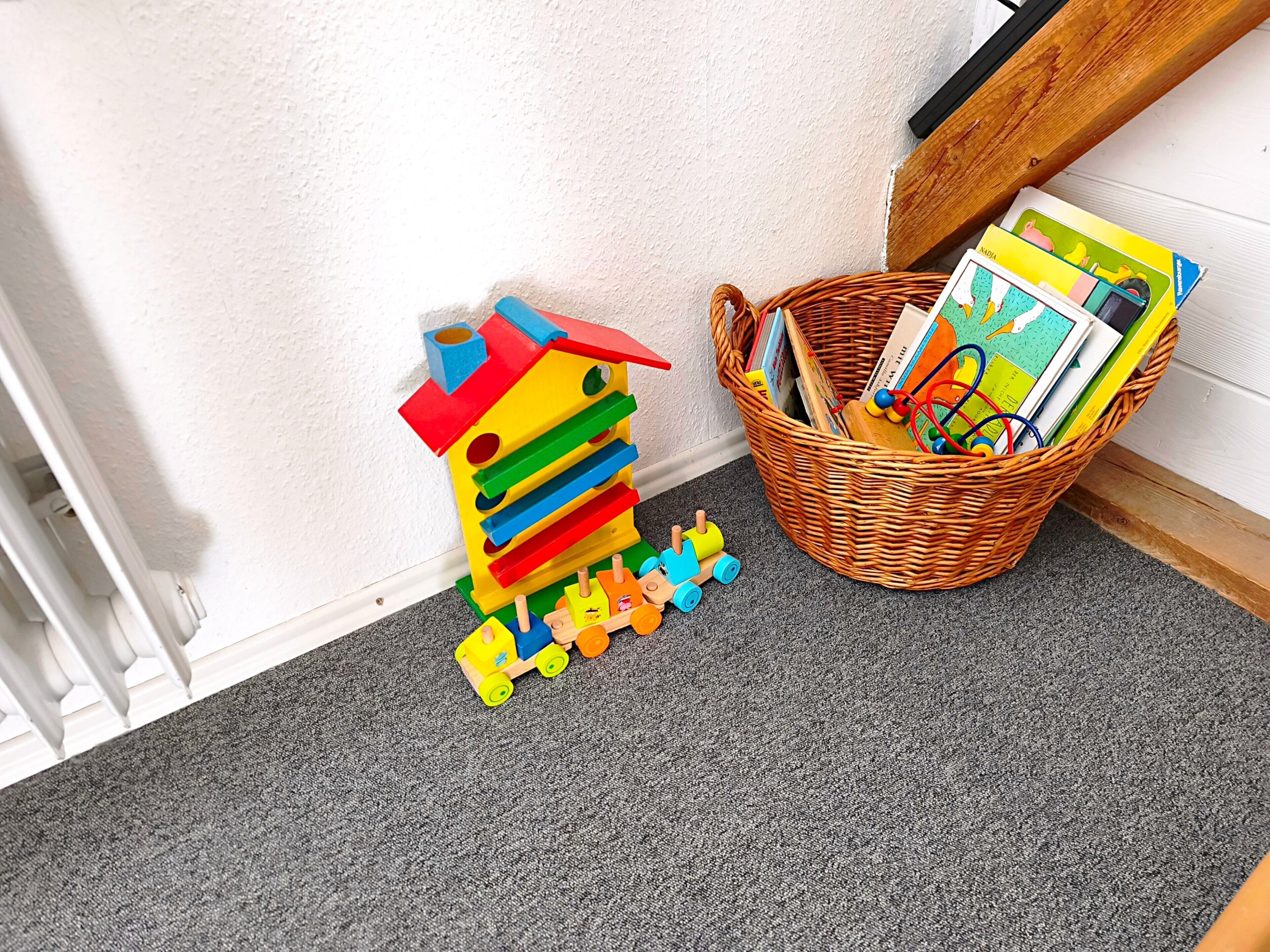 Un coin de pièce avec un mur blanc, une moquette grise, un plancher en bois et un panier en osier rempli de livres et de jouets.