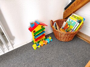 Un coin de pièce avec un mur blanc, une moquette grise, un plancher en bois et un panier en osier rempli de livres et de jouets.