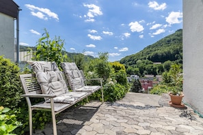 A patio with a cushioned bench, potted plants, and a view of a hillside.