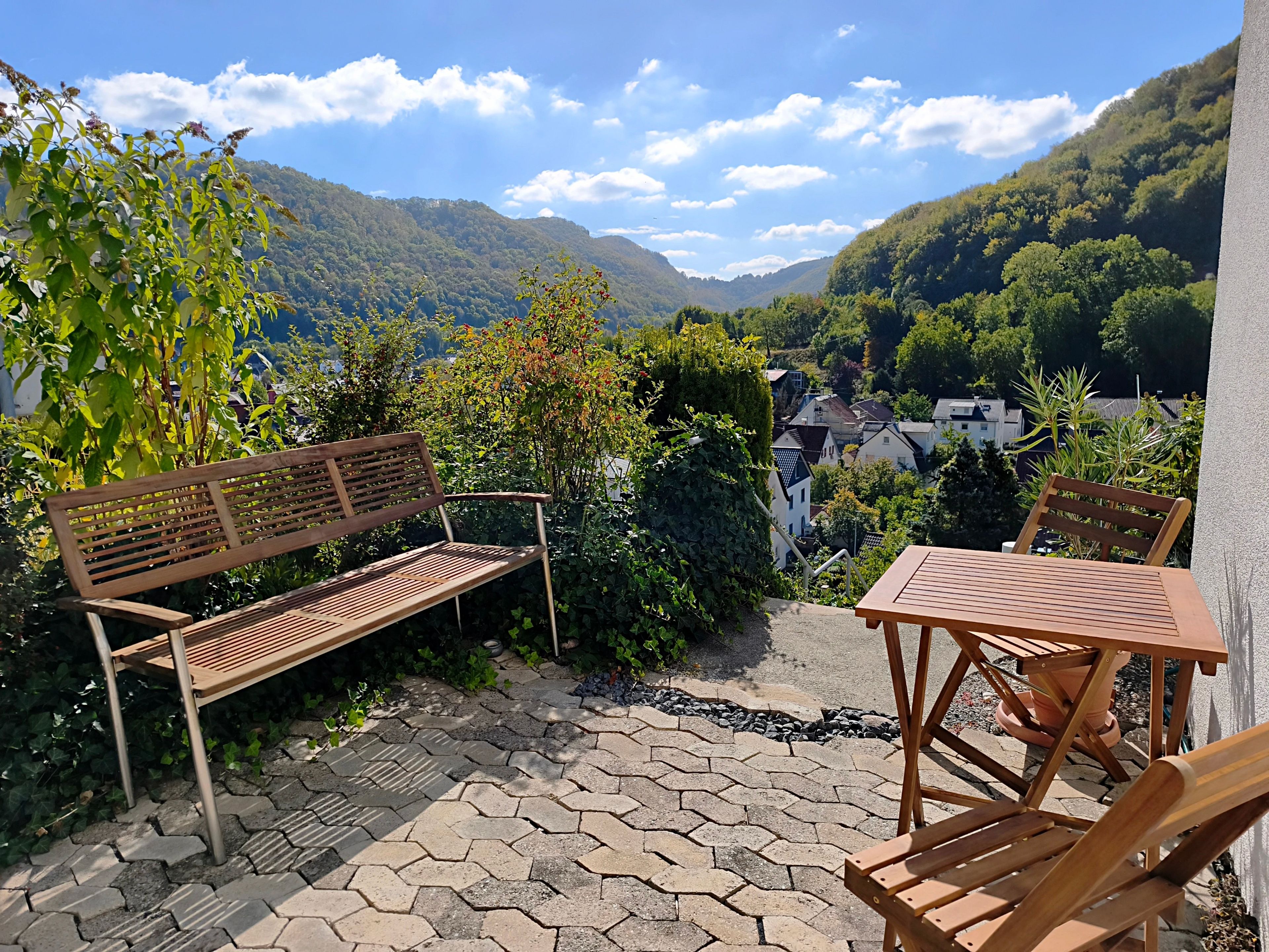 Un patio con un banco y una mesa de madera, rodeado de follaje y vistas a un valle.