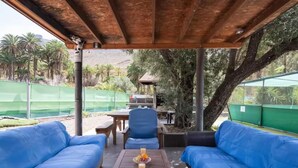 A covered outdoor seating area with blue cushioned sofas, a wooden table with fruit and drinks, and a view of a pool and greenery. - Country Studio Near Fataga (San Bartolomé de Tirajana)