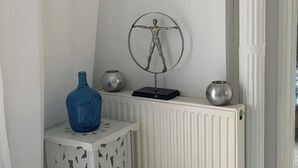 Canto de um quarto com um radiador branco, uma pequena mesa com um vaso azul e uma escultura de metal na parede.