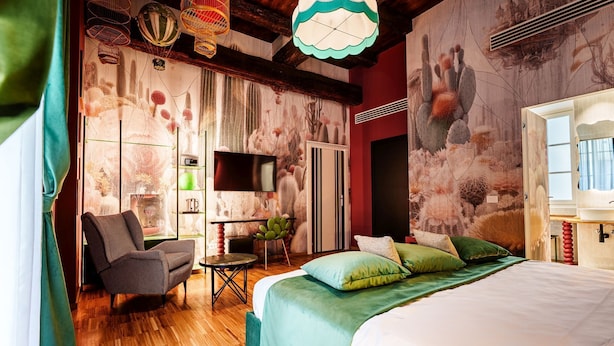 A bedroom with a bed, a chair, a TV, a shelf with items, and a wall with a cactus mural. - Palazzo San Fedele - Como Suites (Como)