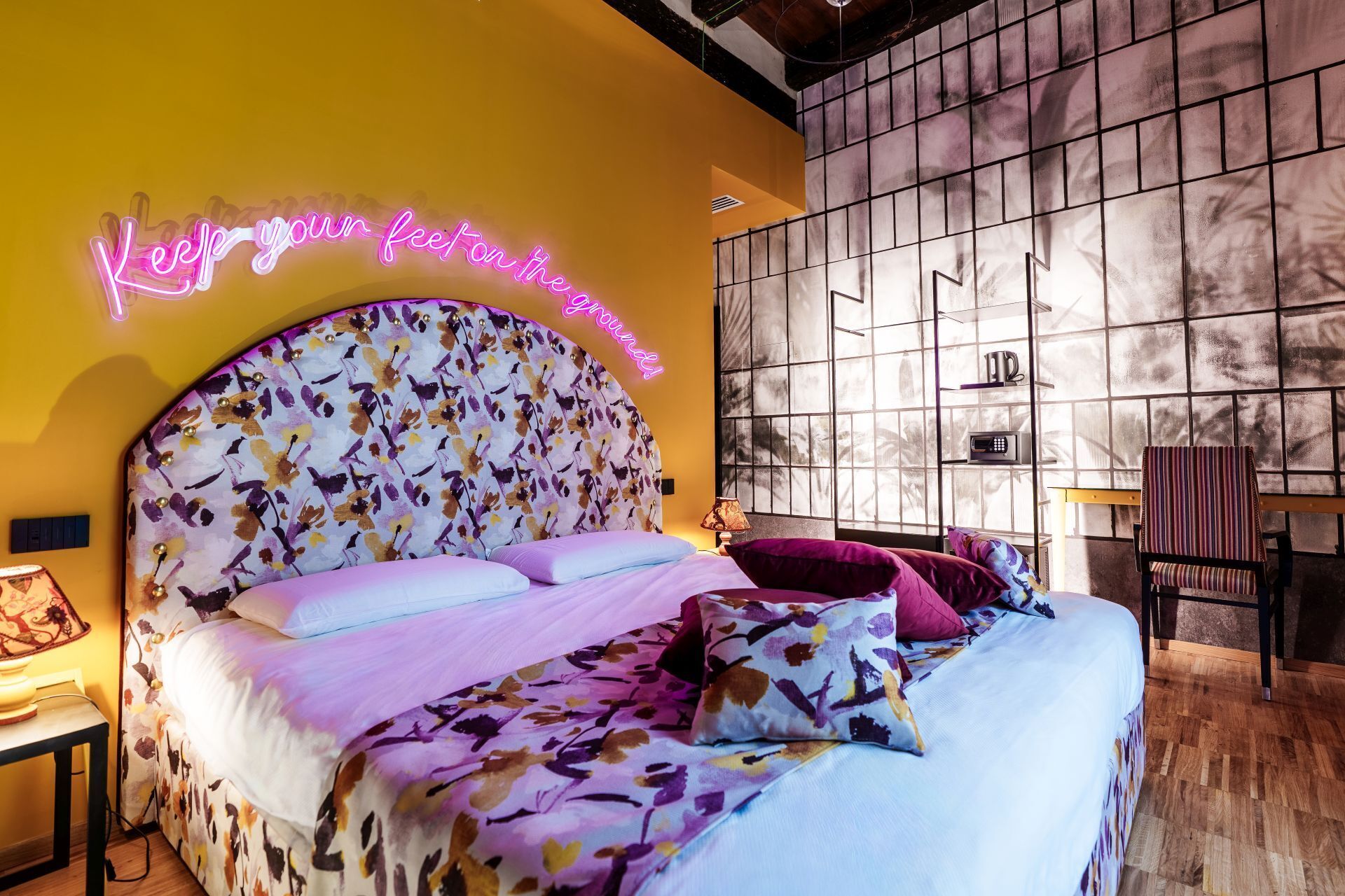 Un dormitorio con cabecera floral, un letrero de neón, una cama con múltiples almohadas, una mesita de noche y una ventana con vistas.
