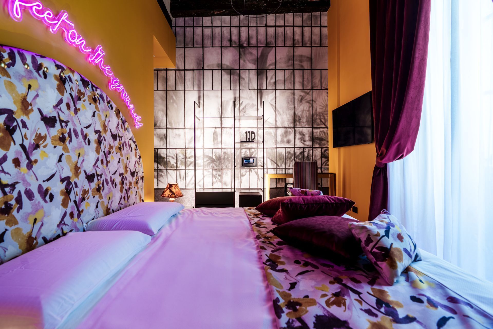 A bedroom with a floral headboard, a bed with purple bedding, a nightstand, a lamp, and a wall-mounted TV.