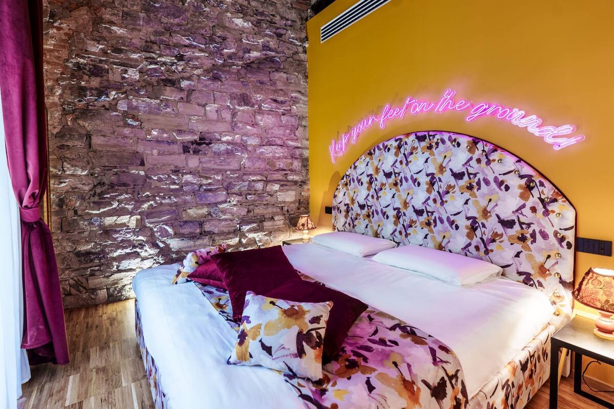 Ein Zimmer mit Backsteinwand, einem Bett mit Blumenmuster am Kopfteil und einem Neonzeichen.