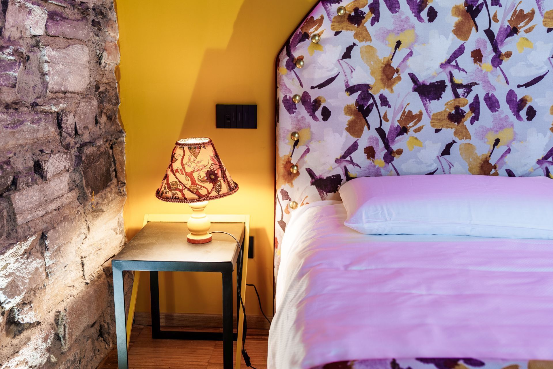 A room with a bed, a lamp on a table, and a brick wall.