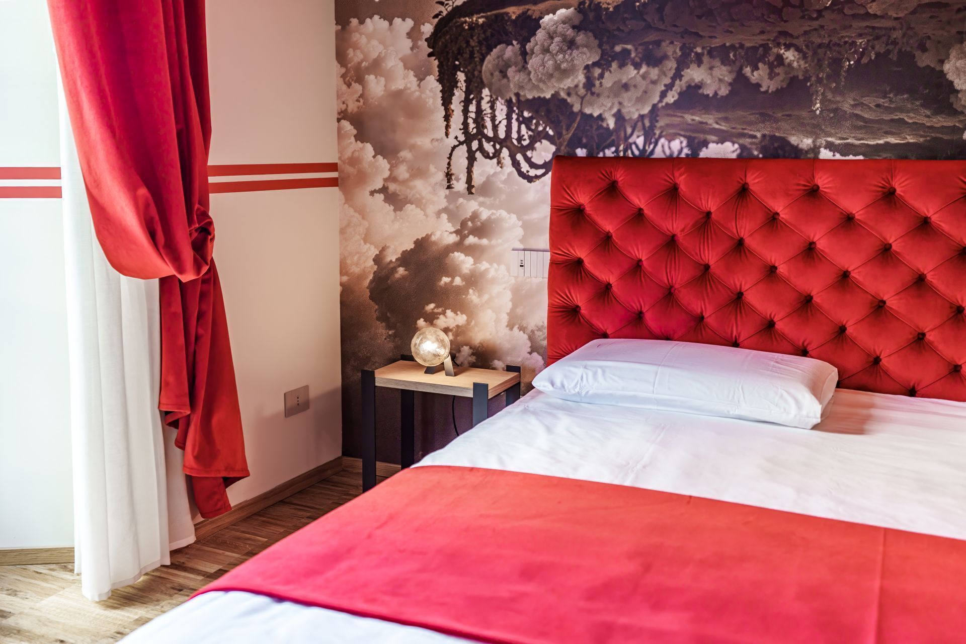 A bedroom with a red tufted headboard, a white bedspread, and a wall mural of clouds.