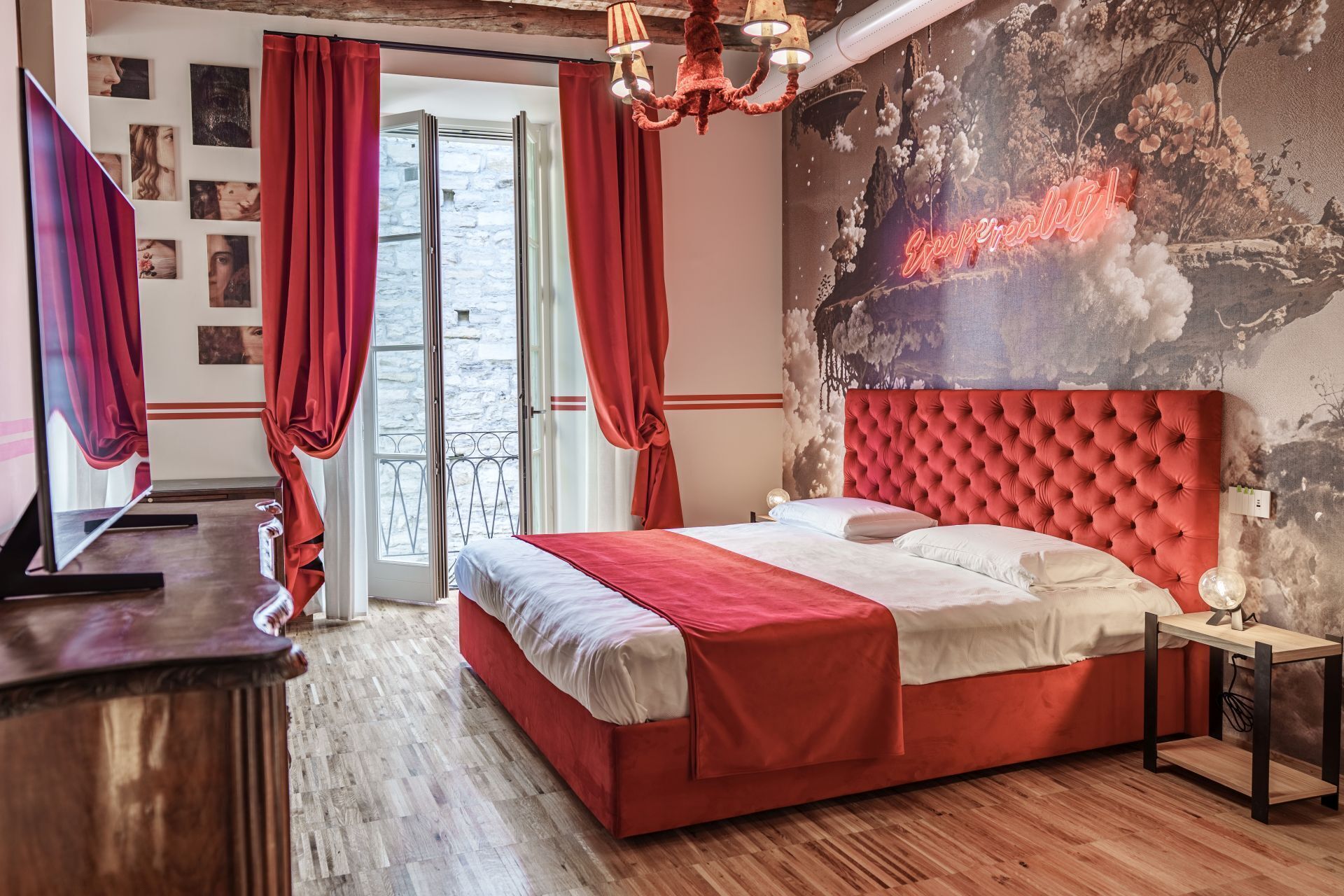 A bedroom with a red bed, a television, a chandelier, and a mural on the wall.